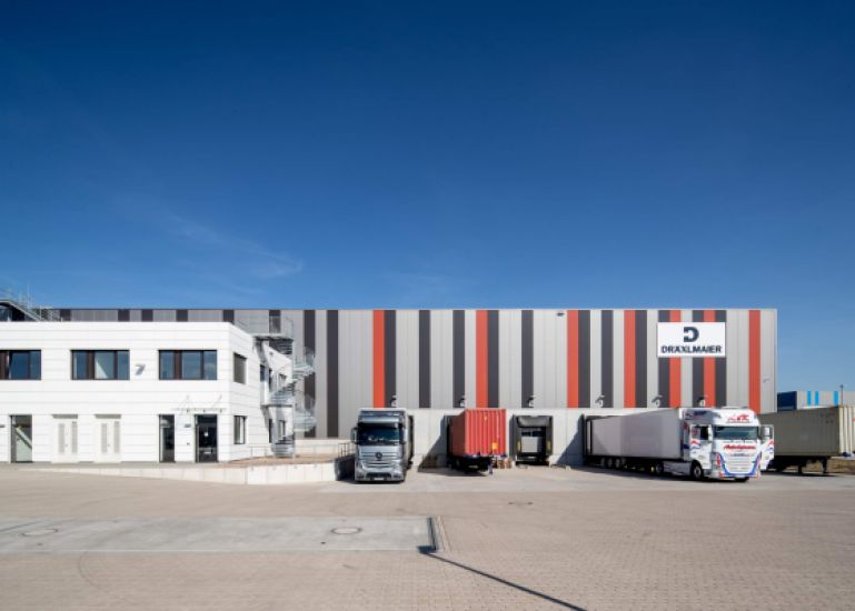 Frontansicht der Lagerhalle Hansalinie Bremen - Dräxlmaier. Zu sehen ist ein Teil des Gebäudes, das teilweise mit farbigen Streifen bemalt ist, davor sind die Luken zum Entladen der LKWs zu sehen, an denen teilweise auch LKWs geparkt sind.