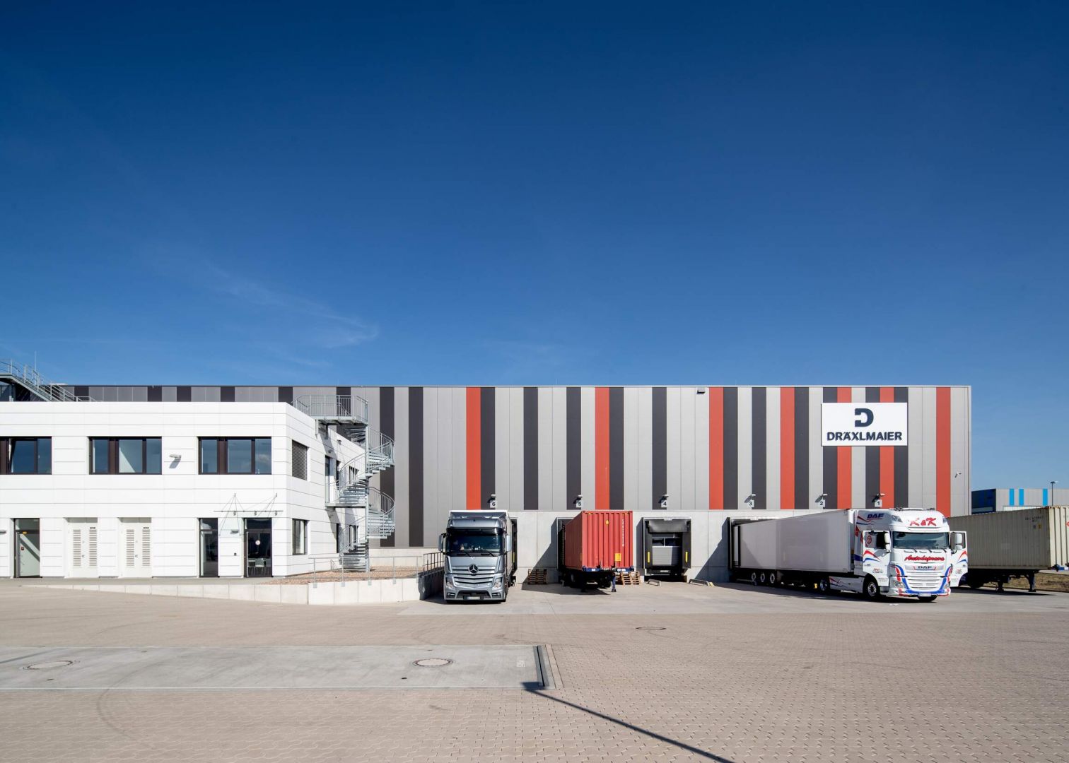 Frontansicht der Lagerhalle Hansalinie Bremen - Dräxlmaier. Zu sehen ist ein Teil des Gebäudes, das teilweise mit farbigen Streifen bemalt ist, davor sind die Luken zum Entladen der LKWs zu sehen, an denen teilweise auch LKWs geparkt sind.