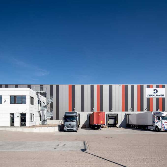 Front view of the Hansalinie Bremen - Dräxlmaier warehouse. You can see part of the building, which is partially painted with coloured stripes, in front of which you can see the hatches for unloading the trucks, some of which are also parked.