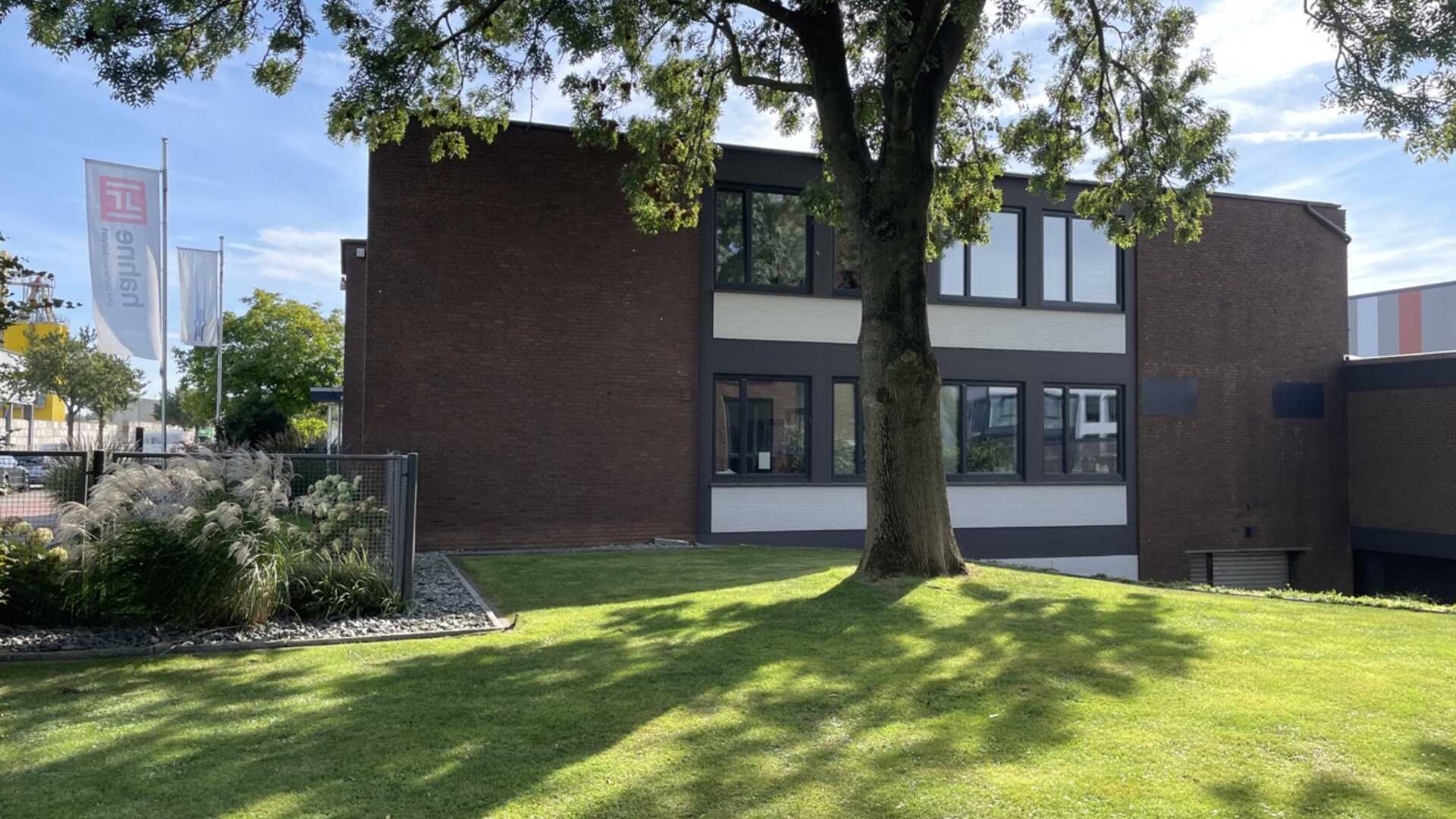 Fotografie eines Bürogebäudes am Allerkai. Zu sehen ist ein Gebäude aus dunklem Stein mit Fensterreihen in der Mitte. Davor eine Grünfläche mit einem Baum.