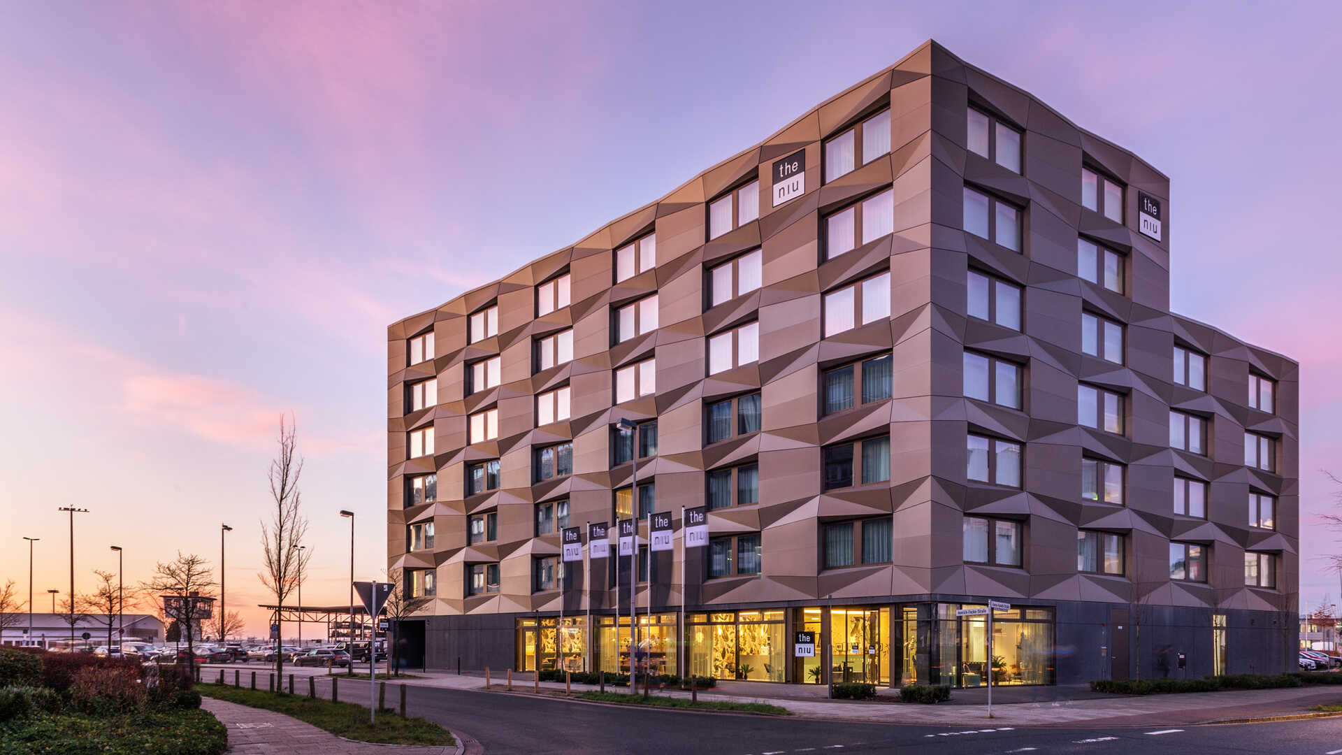 Ein Foto des Hotels "the niu" bei Sonnenuntergang. Es zeigt ein großes Hotel mit eckigen Mustern in der Fassade.  Die untere Etage ist mit einer Glasfront verkleidet und beleuchtet.