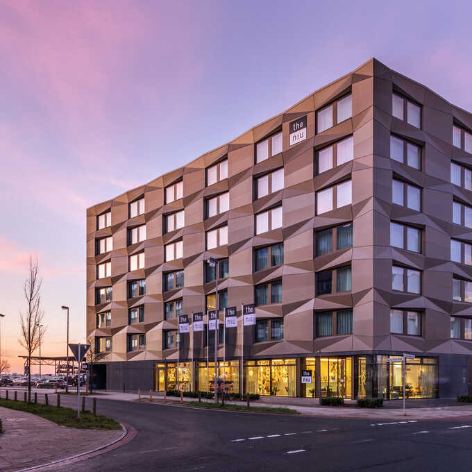 Ein Foto des Hotels "the niu" bei Sonnenuntergang. Es zeigt ein großes Hotel mit eckigen Mustern in der Fassade.  Die untere Etage ist mit einer Glasfront verkleidet und beleuchtet.