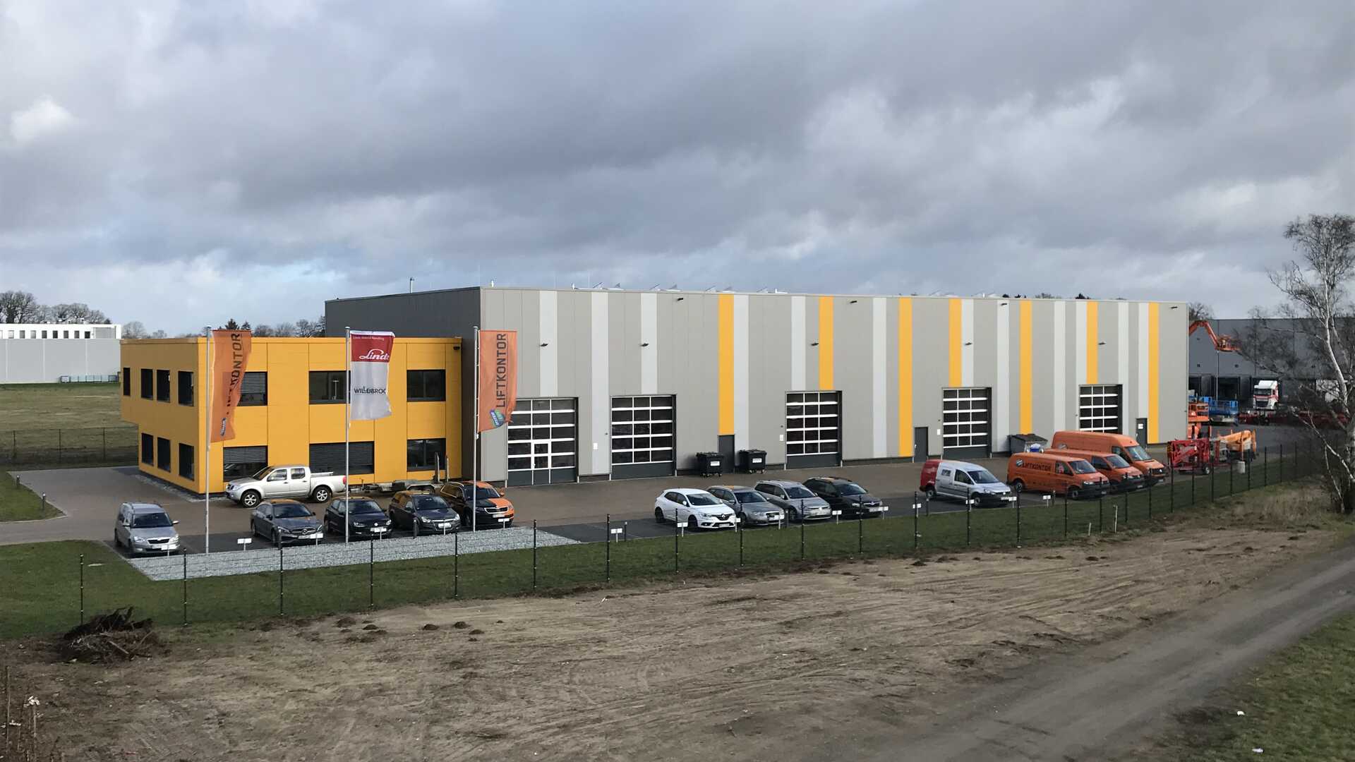 Foto der Lager- und Montagehalle "Liftkontor Hannover". Abgebildet ist eine große Halle mit mehreren LKW-großen, halbverglasten Garagen. Davor befindet sich ein Parkplatz, auf dem Pkw abgestellt sind.