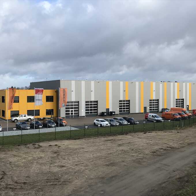 Foto der Lager- und Montagehalle "Liftkontor Hannover". Abgebildet ist eine große Halle mit mehreren LKW-großen, halbverglasten Garagen. Davor befindet sich ein Parkplatz, auf dem Pkw abgestellt sind.