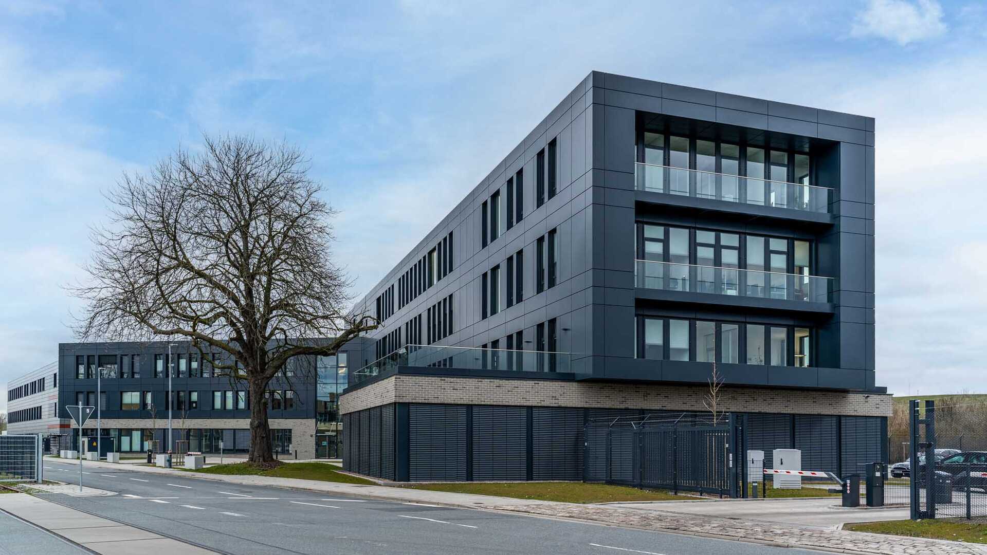 Foto des Bürogebäudes "ADAS". Dargestellt ist ein großes, dunkles Gebäude mit schmalen, bodentiefen Fenstern und einer durchgehenden Fensterfront auf einer Seite. Vor dem Gebäude befindet sich eine Zufahrt zu Parkplätzen.