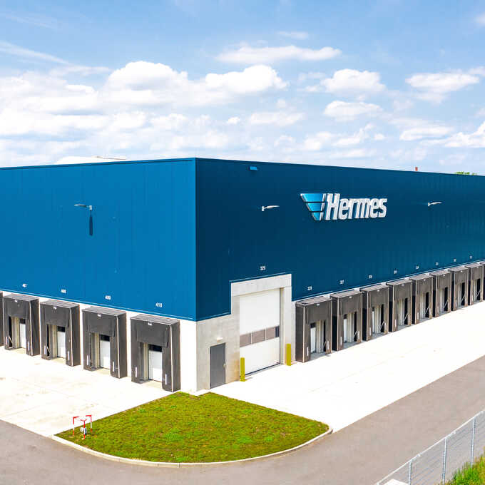 Foto des Hochleistungslagers Weyhe - Hermes. Abgebildet ist eine große Warenhalle mit reihenweise Verlademöglichkeiten für Transporter und LKW. Oben das Logo der Firma Hermes mit dem Schriftzug "Hermes".